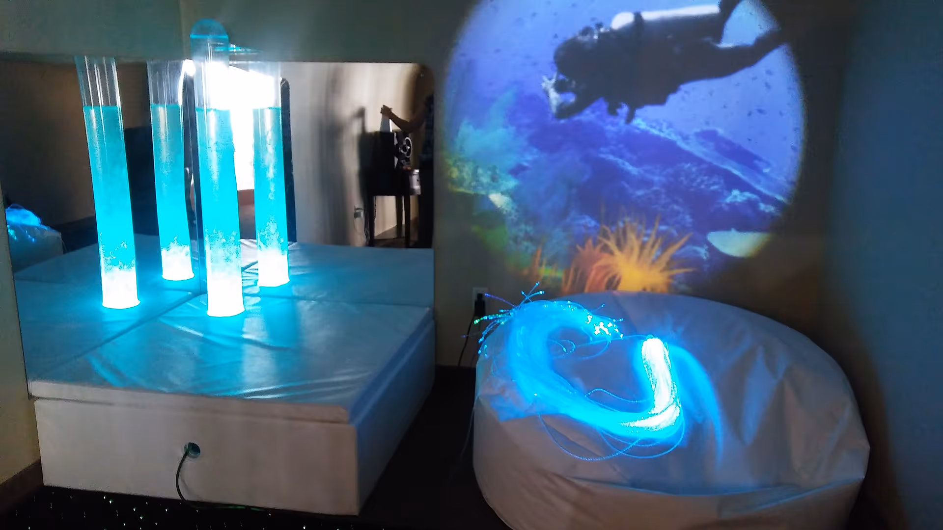 A sensory room with illuminated bubble tubes on a padded platform next to a large mirror, a white bean bag chair with glowing fiber optic strands on it, and a projection of an underwater scene with a scuba diver and coral on the wall.