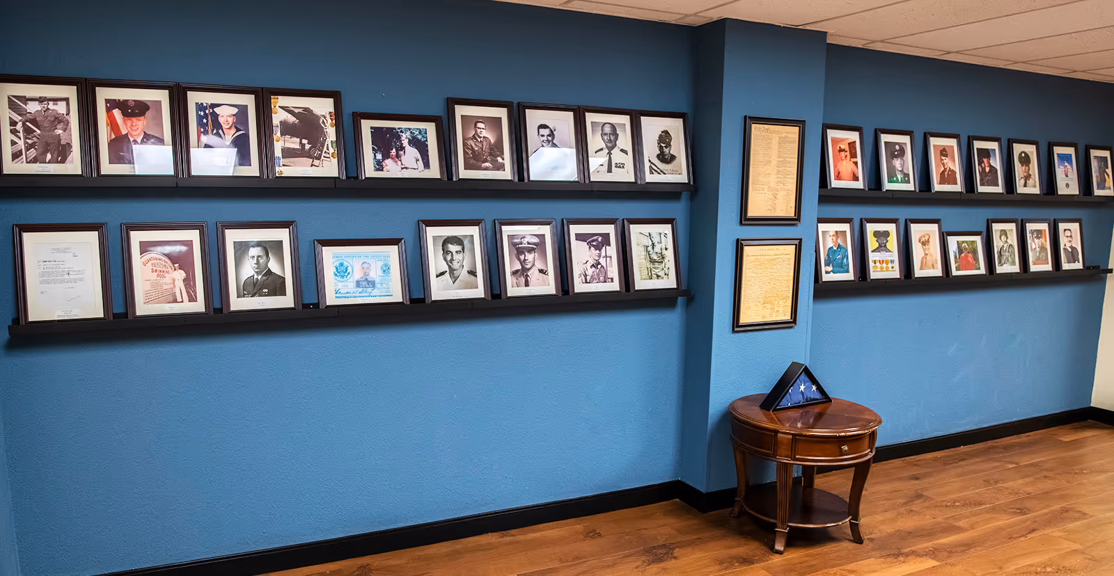 Blue-walled interior with two long shelves of framed photos and documents and a small round wooden table holding a folded flag.