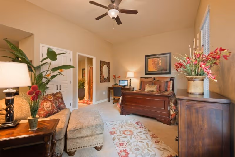 A cozy bedroom with a wooden bed featuring patterned bedding, a wooden dresser with a vase of red flowers, a small desk with a chair and a lamp, and a beige sofa with a matching ottoman. The room has beige walls, a ceiling fan, a floral rug, and a large plant near a side table with a lamp.