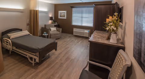 A spacious, well-lit rehabilitation room with a hospital bed on the left, a comfortable armchair with a side table and lamp near the window, a large wooden dresser with a vase of flowers and a TV on top, and a chair in the foreground. The room has wood flooring and neutral-colored walls.