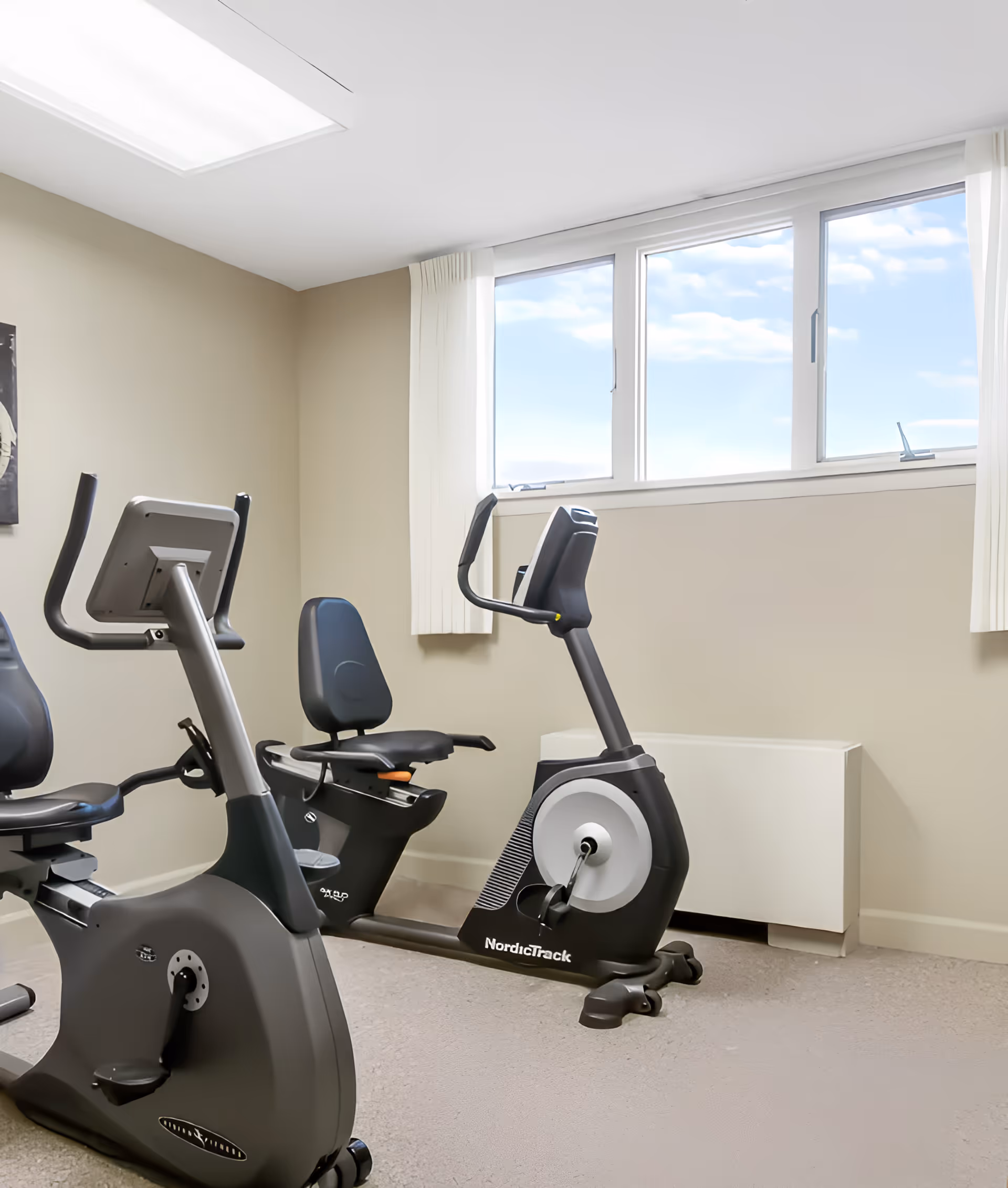 A small exercise room with two stationary recumbent bikes positioned near a window with white curtains. The walls are beige, and the floor is carpeted. The room is well-lit with natural light coming through the window.