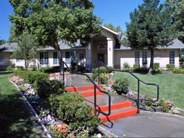 Exterior view of a single-story building with a walkway and red steps leading to the entrance. The building is surrounded by green grass, trees, and colorful flower beds under a clear blue sky.