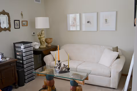 A cozy living room with a white patterned sofa against a beige wall, decorated with three framed artworks. In front of the sofa is a glass coffee table with a unique wooden base and three candles in holders. To the left, there is a wooden side table with a lamp, a decorative statue, and a stack of plastic drawers. Various small framed pictures and a mirror hang on the wall.