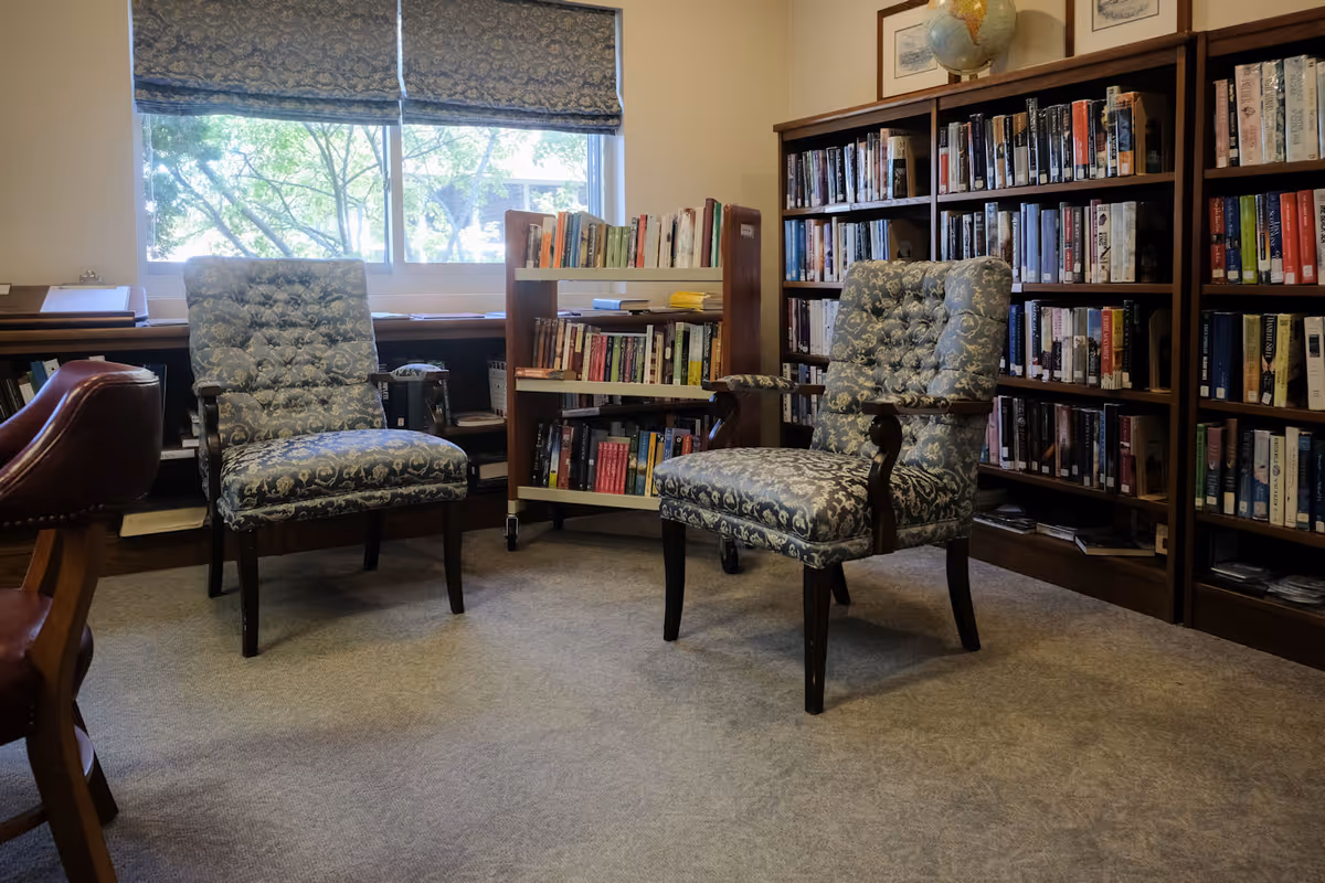A cozy library room with two patterned upholstered armchairs placed on a carpeted floor. Behind the chairs are bookshelves filled with books and a rolling book cart. A window with a patterned Roman shade lets in natural light, and a globe is visible on top of one bookshelf.