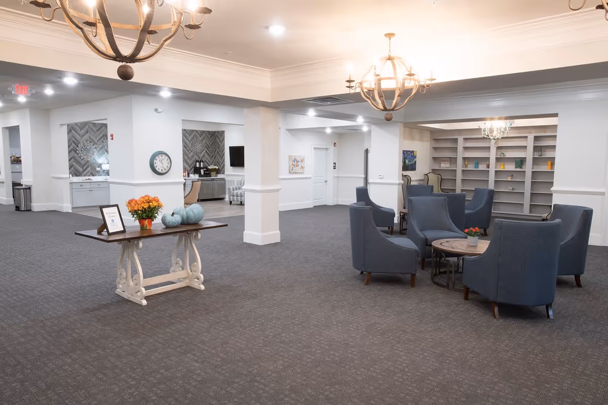 A spacious and well-lit common area in a senior living facility featuring a seating arrangement with six blue armchairs around a round wooden coffee table with a small potted plant. The room has gray carpet flooring, white walls with molding, and built-in shelves with decorative items. Two large chandeliers hang from the ceiling, and a wooden table with flowers and decorative pumpkins is placed near the center. The background shows a kitchenette area with a clock on the wall and a TV.