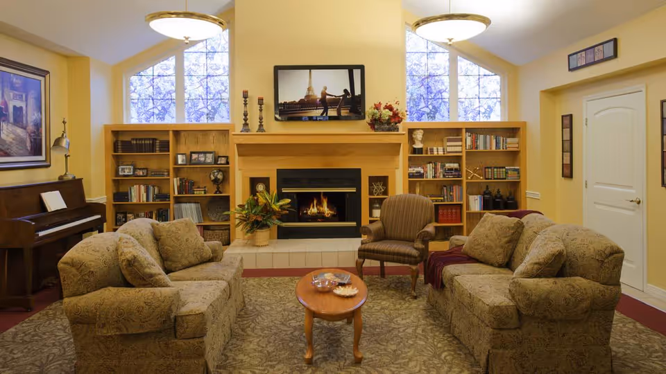 A cozy living room with two patterned sofas facing each other and a single striped armchair. A wooden coffee table sits in the center on a patterned rug. Behind the seating area is a fireplace with a fire burning, flanked by two wooden bookshelves filled with books and decorative items. Above the fireplace is a flat-screen TV displaying an image of the Eiffel Tower. Large windows with a view of tree branches are on either side of the fireplace. A piano with a lamp and framed artwork is on the left side of the room. The walls are painted yellow and the room is warmly lit.