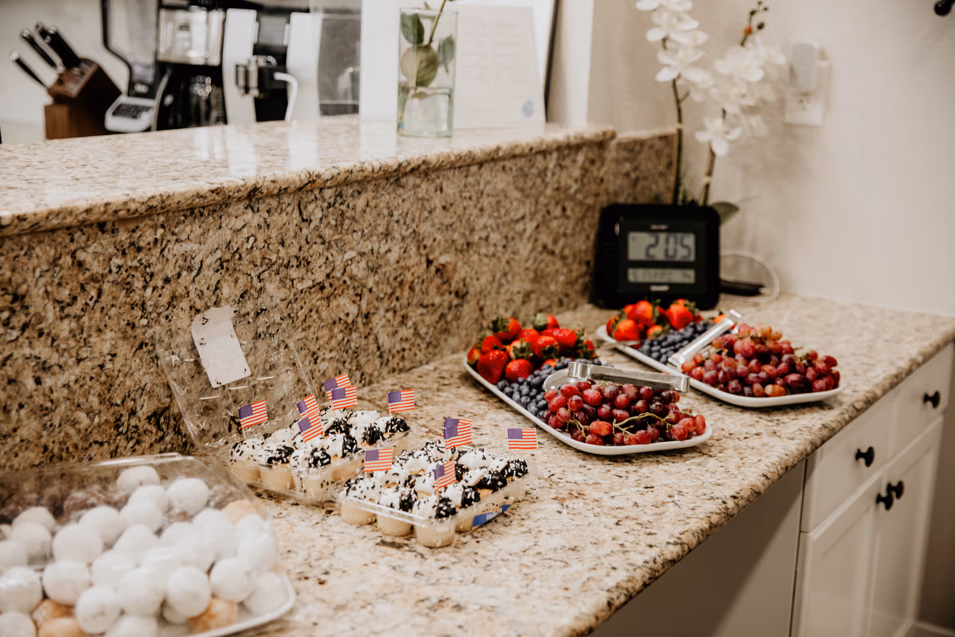 A kitchen countertop with plates of fresh fruit including strawberries, blueberries, and grapes, alongside trays of small desserts topped with miniature American flags. In the background, there is a coffee maker, a knife block, a digital clock, and a vase with white flowers.
