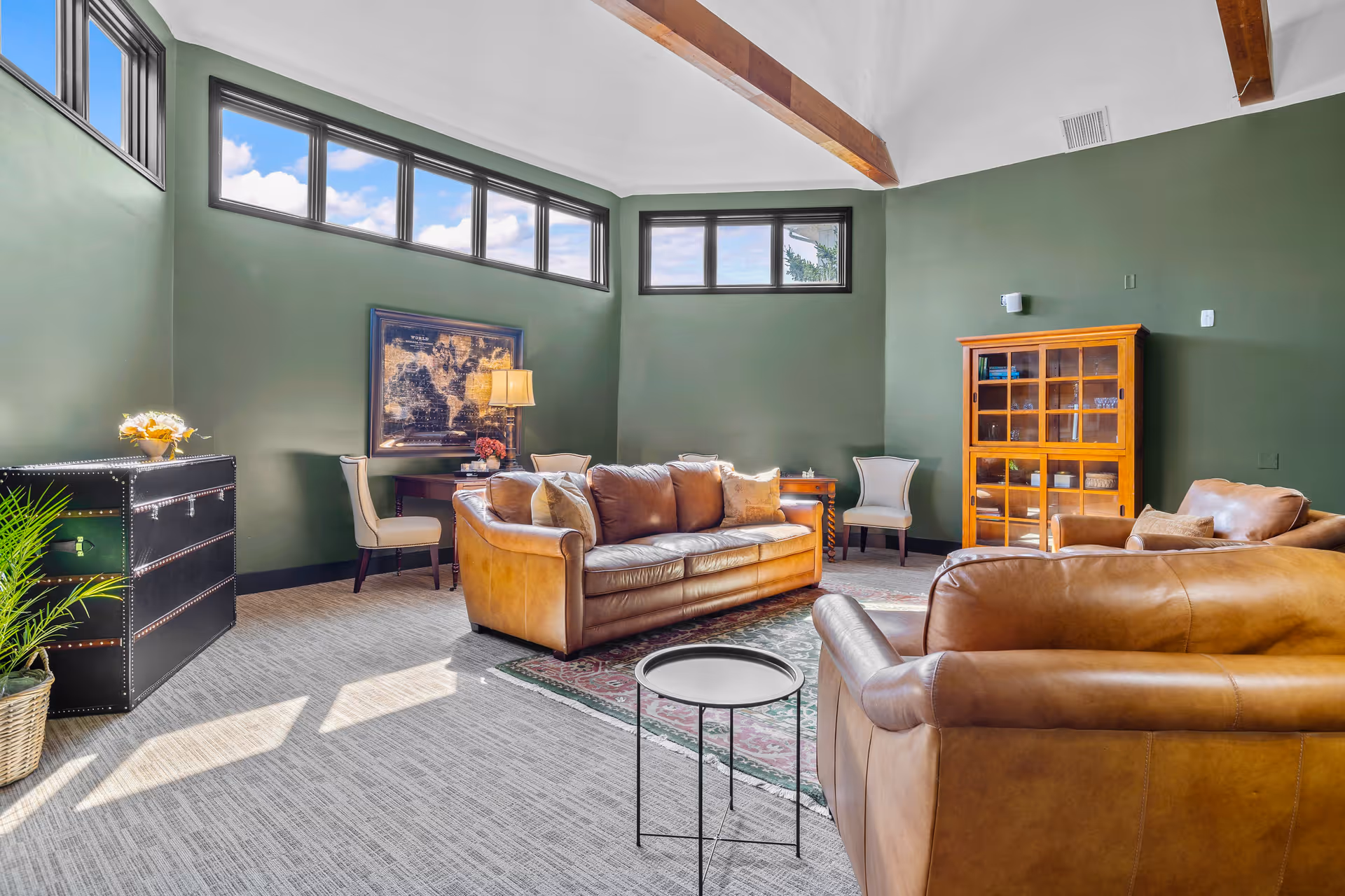 A bright living room with green walls and high windows letting in natural light. The room features brown leather sofas, a small round side table, a wooden cabinet with glass doors, two upholstered chairs, a black chest, a potted plant, and a framed map on the wall. The ceiling has exposed wooden beams.