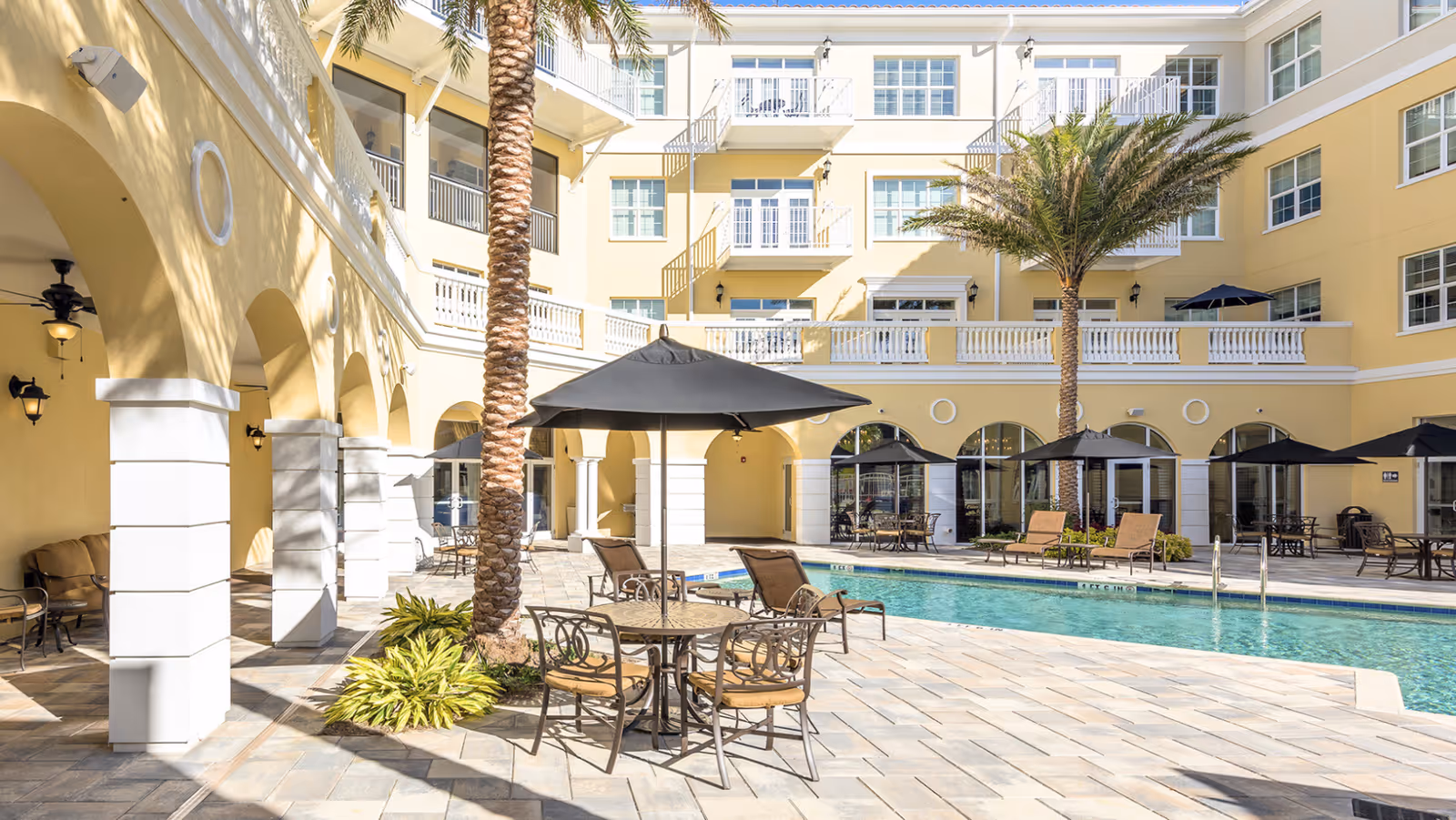 Outdoor courtyard area of a senior living facility with a swimming pool, palm trees, patio tables with umbrellas, lounge chairs, and a multi-story yellow building with balconies and arched walkways surrounding the pool.