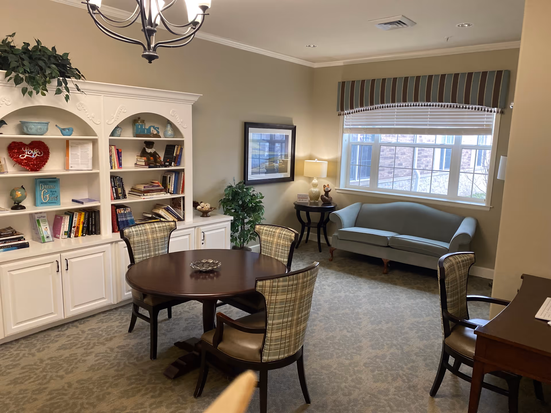 A cozy sitting area in a senior living facility featuring a round wooden table surrounded by four plaid upholstered chairs. Behind the table is a white built-in bookshelf filled with books and decorative items. To the right, there is a light blue upholstered sofa beneath a window with striped valance and blinds. A small side table with a lamp and decorative items is next to the sofa. The room has beige walls, carpeted floor, and a chandelier hanging from the ceiling.