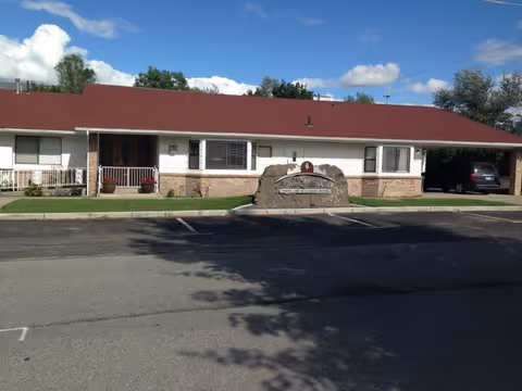 Single-story building with a red roof and white walls, featuring a front porch with railings and a large stone sign in front that reads 'Aspen Quality Care Inc.'. There is a parking area in front of the building and a car parked under a carport on the right side. Trees and a blue sky with clouds are visible in the background.