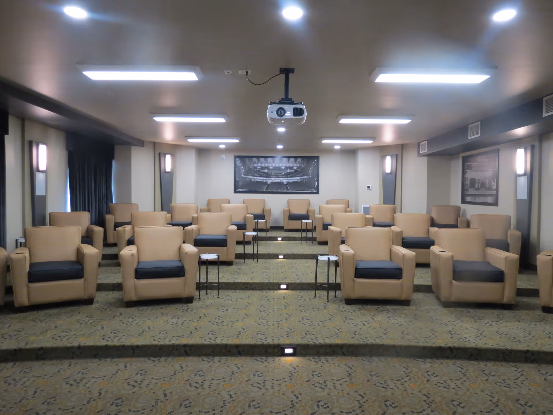 Tiered media room with rows of beige armchairs facing a ceiling-mounted projector and screen.