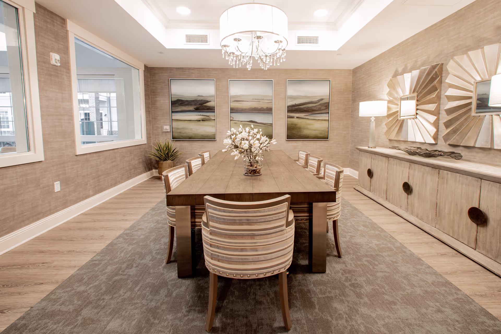 A modern dining room with a long wooden table surrounded by eight striped upholstered chairs. The room features a large chandelier hanging above the table, three landscape paintings on the far wall, a sideboard with decorative mirrors and a lamp, and a potted plant in the corner. The floor has a light wood finish with a large area rug under the table.