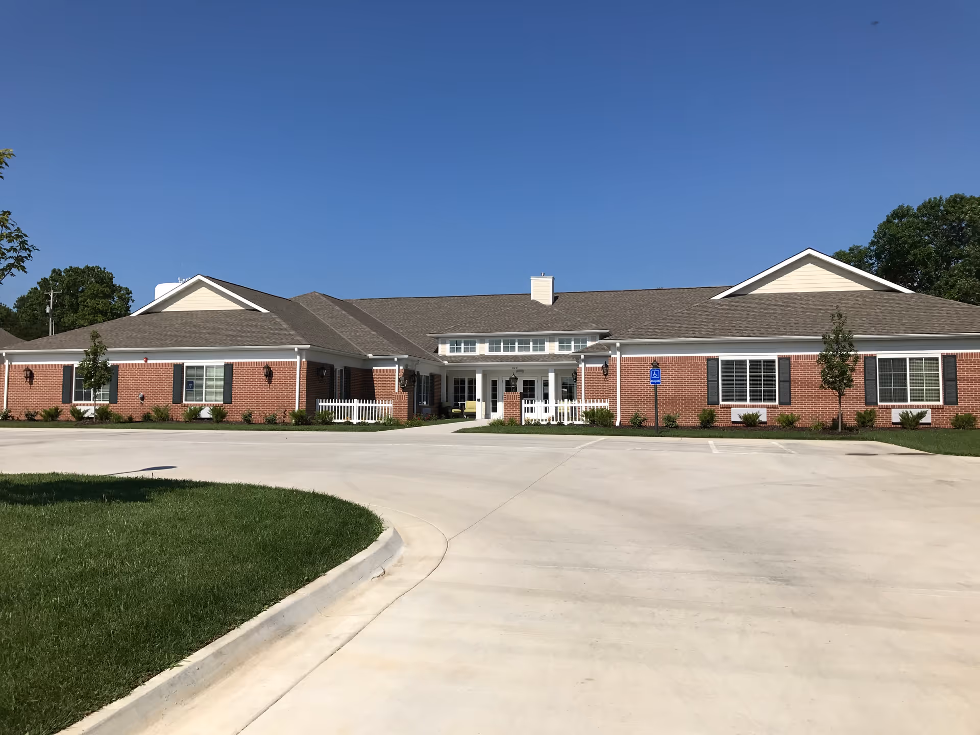 Front exterior of a single-story brick senior living building with a wide concrete driveway, small landscaped beds, and a clear blue sky.