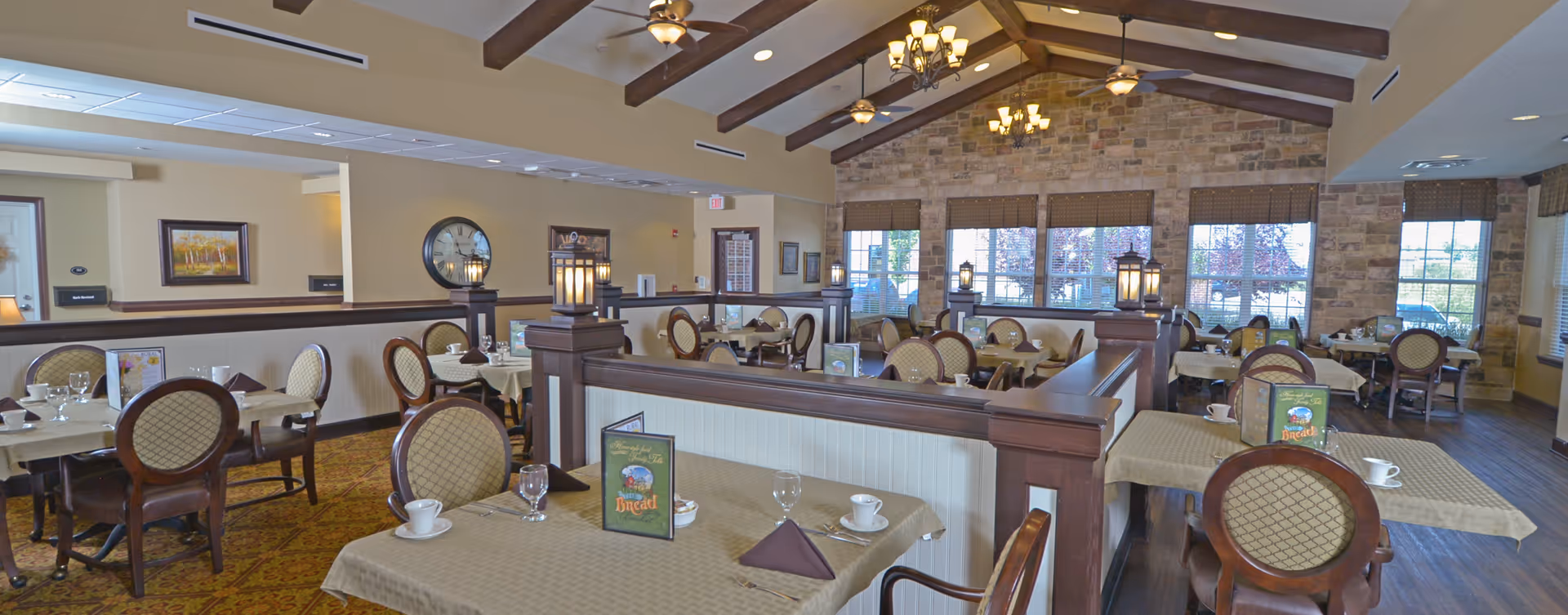 A spacious dining room with multiple tables covered in beige tablecloths, each set with cups, glasses, napkins, and menus. The room features wooden chairs with cushioned seats and backs, a high ceiling with exposed wooden beams, ceiling fans, and chandeliers. Large windows with blinds allow natural light to fill the room, and the walls are decorated with stone and framed artwork.