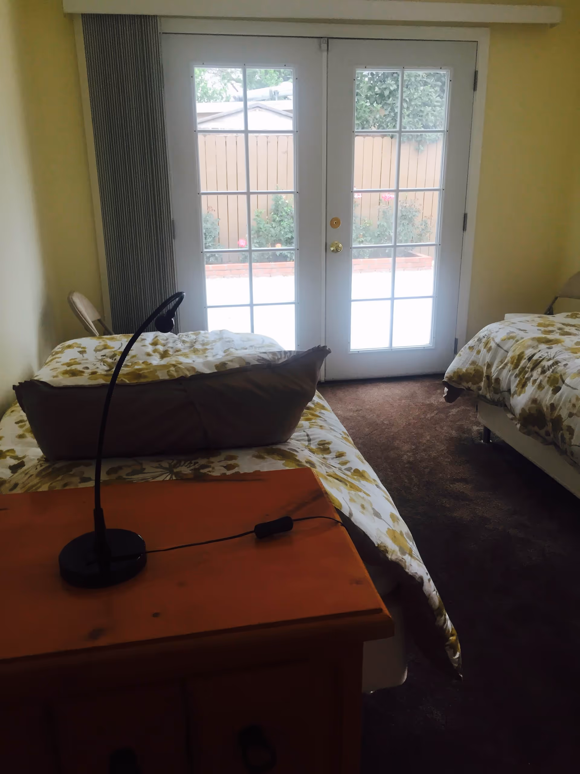 A bedroom with two single beds covered in floral-patterned bedding. A wooden nightstand with a black reading lamp is positioned between the beds. The room has yellow walls and a brown carpet. At the far end, there are white French doors with glass panes leading to an outdoor area with a wooden fence and some plants.
