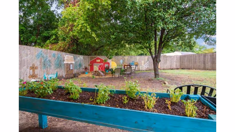 Outdoor garden area with a raised blue planter box containing small plants in the foreground. In the background, there is a fenced yard with a tree, several chairs around a fire pit, and a small red playhouse. The wooden fence has colorful murals painted on it.