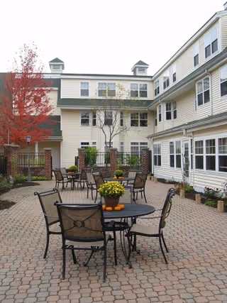 Outdoor paved courtyard with round patio tables and chairs surrounded by a multi-story beige senior living building.