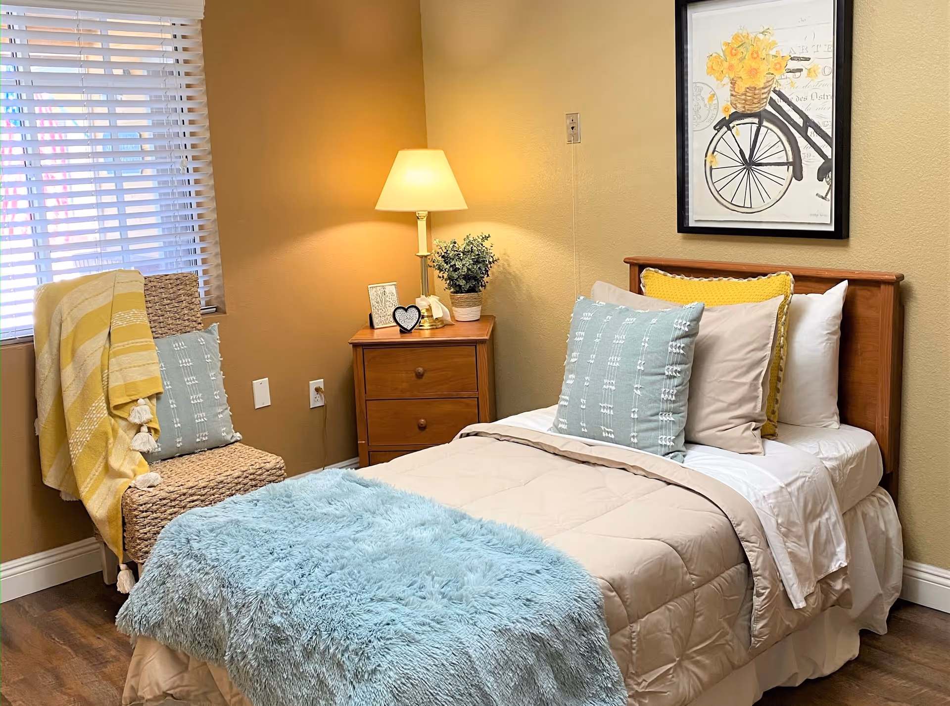 A cozy bedroom with a single bed made with beige and white bedding, multiple pillows including a blue textured one, and a light blue fluffy throw blanket. Next to the bed is a wooden nightstand with a lamp, a small plant, and decorative items. A wicker chair with a yellow and white striped blanket and a blue pillow is placed near a window with white blinds. A framed picture of a bicycle with a basket of yellow flowers hangs on the wall above the bed.