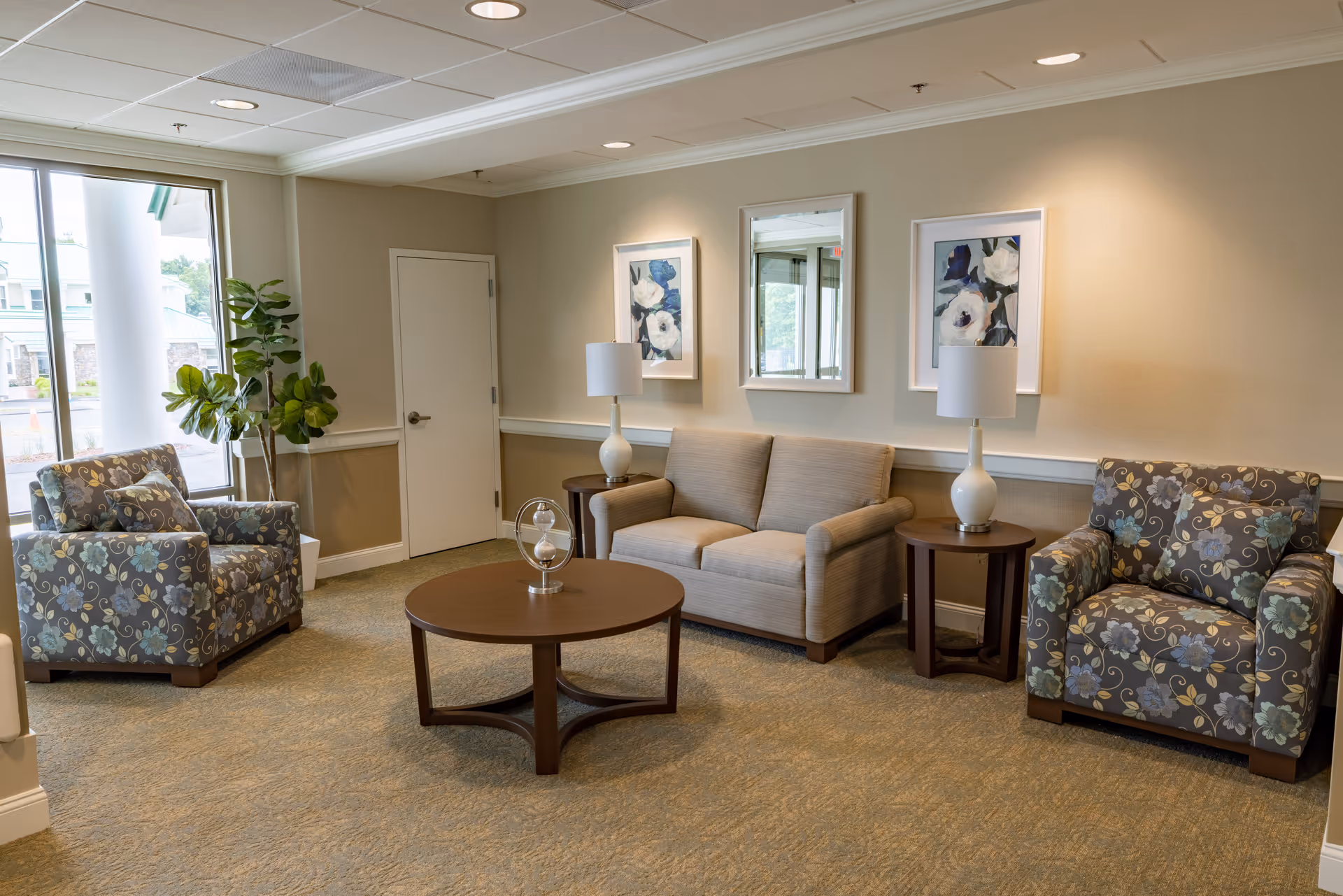 A cozy living room area with a beige loveseat, two floral patterned armchairs, a round wooden coffee table with a decorative hourglass, two side tables each with a white lamp, two floral paintings, a mirror on the wall, and a large window showing an exterior view.