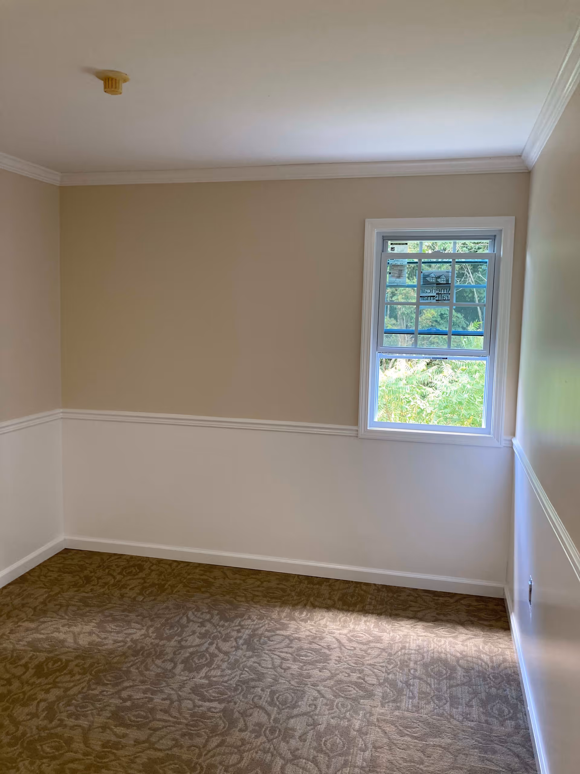 Empty room with beige and white walls, a small window showing greenery outside, patterned carpet flooring, and a smoke detector on the ceiling.