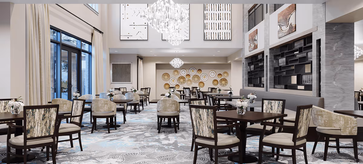 A spacious and elegant dining room with multiple dark wood tables and chairs featuring patterned upholstery. The room has large windows with beige curtains, modern chandeliers hanging from a high ceiling, and decorative wall art including circular plates and abstract paintings. The floor is covered with a patterned carpet in shades of gray and blue.