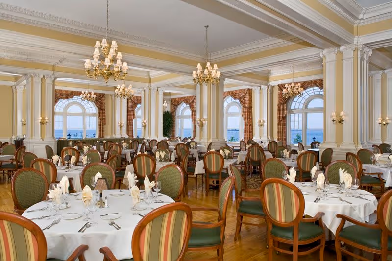 Elegant dining room with round tables covered in white tablecloths, set with folded napkins, silverware, and glassware. The room features large arched windows with views of water outside, ornate chandeliers hanging from a coffered ceiling, and classic wooden chairs with green and striped upholstery.