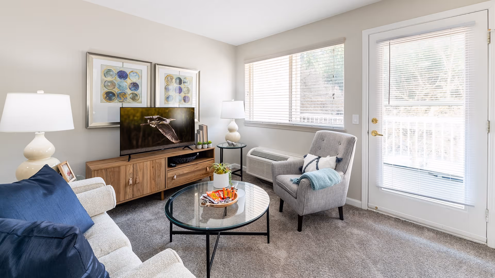 A bright living room with a beige sofa adorned with blue pillows, a gray armchair with a white and blue pillow and a blue throw, a round glass coffee table with a small plant and snacks, a wooden TV stand with a flat-screen TV displaying a bird in flight, two framed abstract artworks on the wall, two white table lamps, a large window with blinds, and a glass door leading outside.