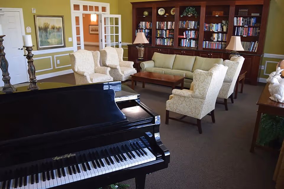 A cozy living room area with a black grand piano in the foreground, cream-colored armchairs, a light green sofa, wooden coffee table, and a large wooden bookshelf filled with books and decorative items against a green wall.