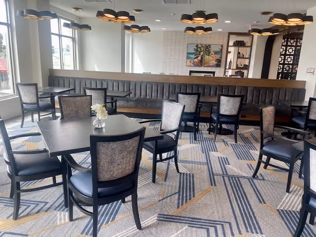A bright dining area with multiple black tables and chairs arranged on a patterned carpet. There is a long cushioned bench along the back wall, large windows letting in natural light, modern ceiling lights, and a decorative shelving unit with artwork and plants.