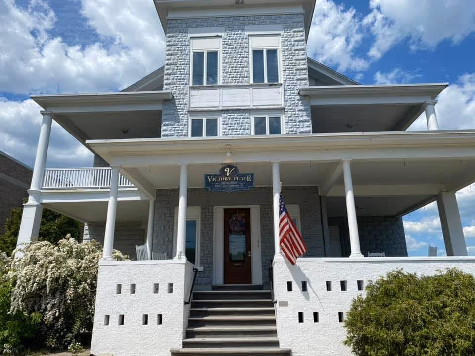 Front exterior view of a multi-story building named Victory Place with a large porch supported by white columns, an American flag displayed near the entrance, and bushes on either side of the stairs leading up to the door. The sky is partly cloudy.