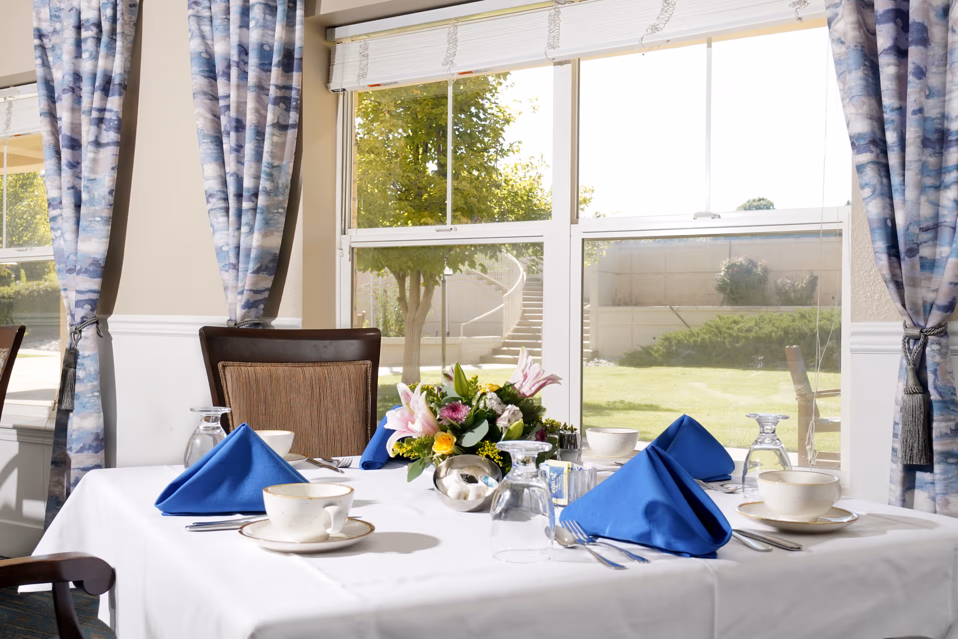 Dining table set with blue napkins, teacups, glasses and a floral centerpiece in front of large windows with blue-patterned curtains.