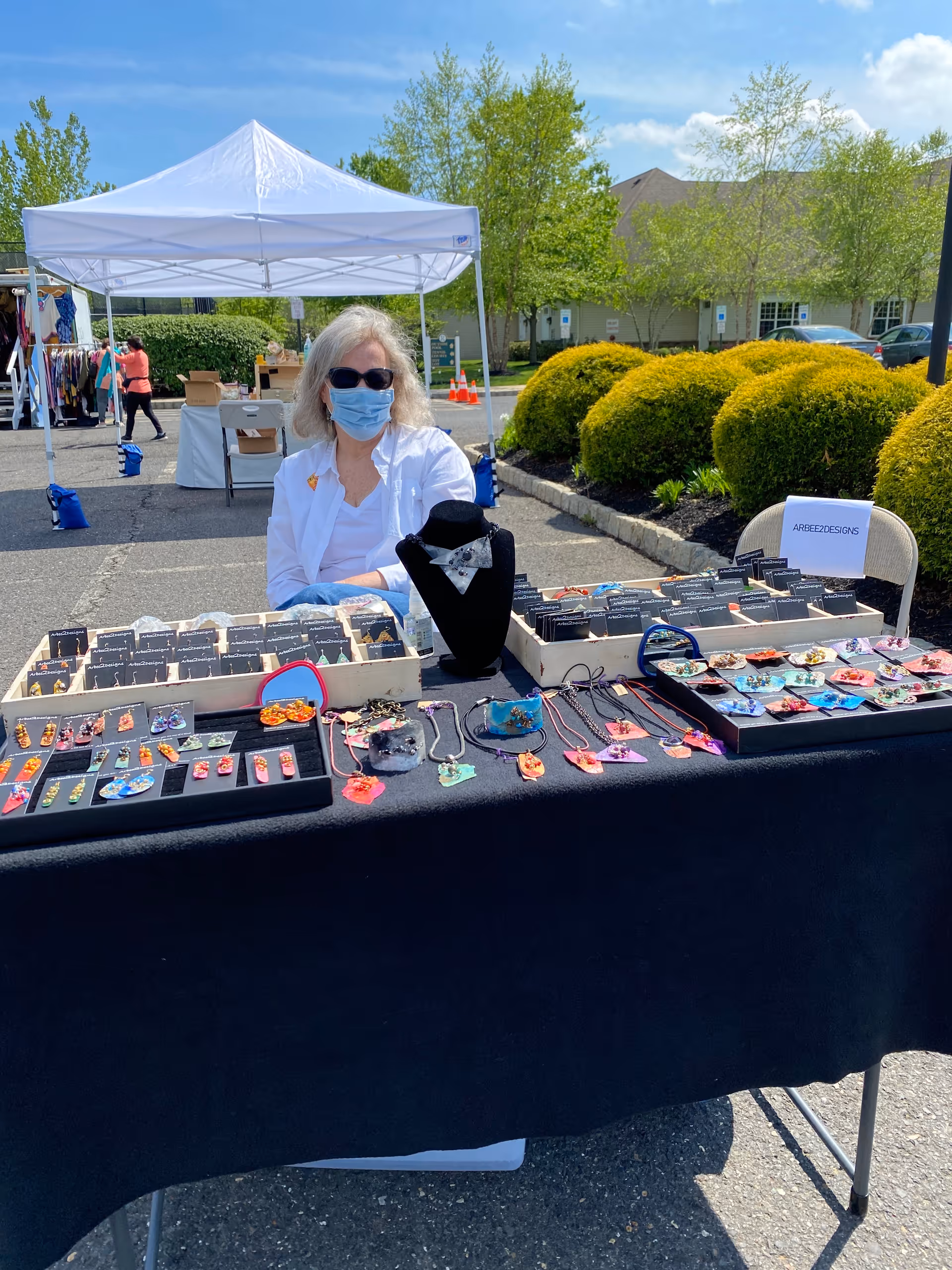 An outdoor market stall with a woman wearing sunglasses and a face mask sitting behind a table displaying various handmade jewelry items including earrings, necklaces, and bracelets. The stall is set up under a white canopy tent with green bushes and a building in the background on a sunny day.