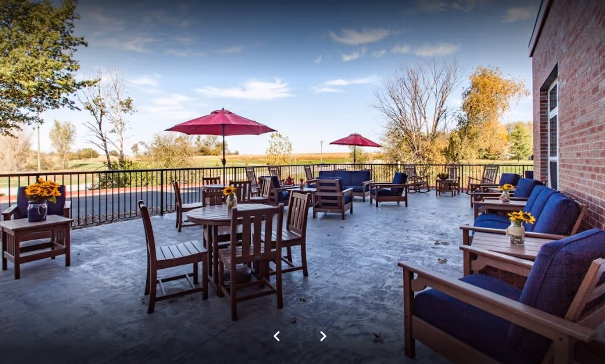 Outdoor patio area with wooden tables and chairs, some with blue cushions, and two red umbrellas. The patio overlooks a scenic view with trees and open sky.
