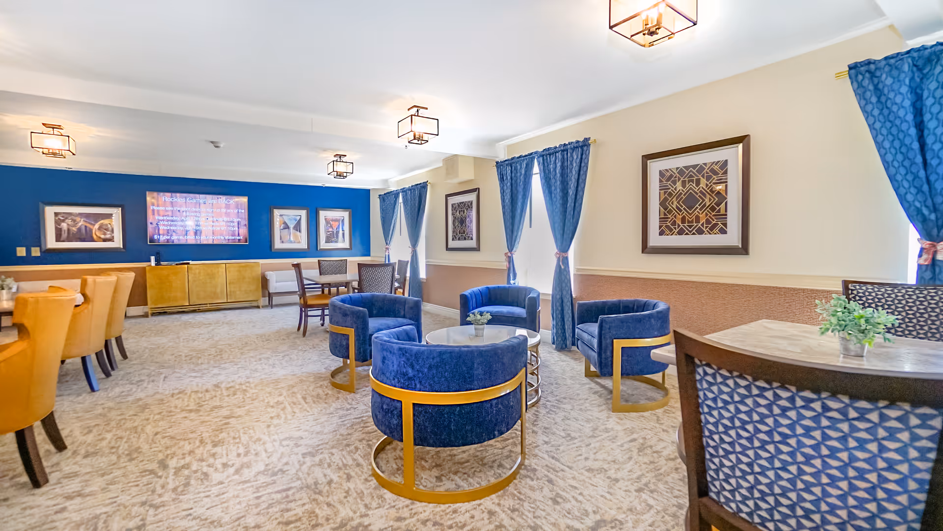 Bright communal lounge with blue velvet chairs around round tables, dining seating, and a wall-mounted TV.