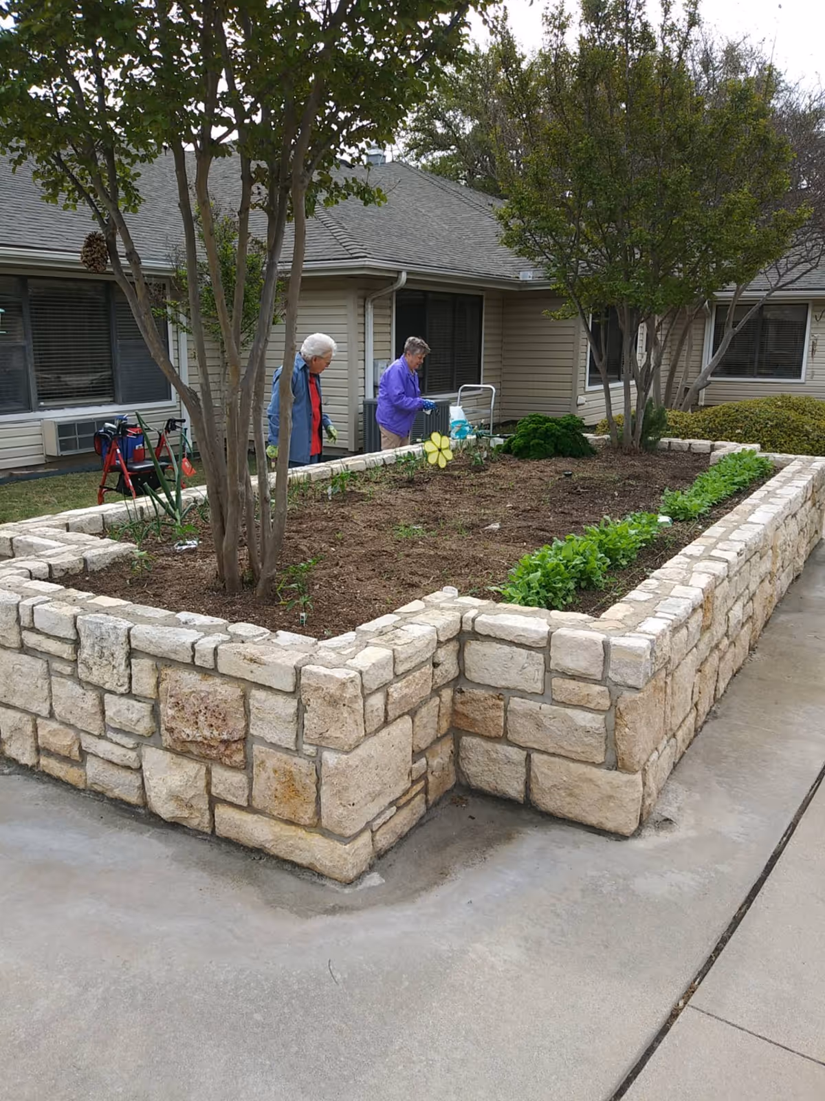 Two elderly women gardening by a raised stone planter in the courtyard of a single-story senior living building.