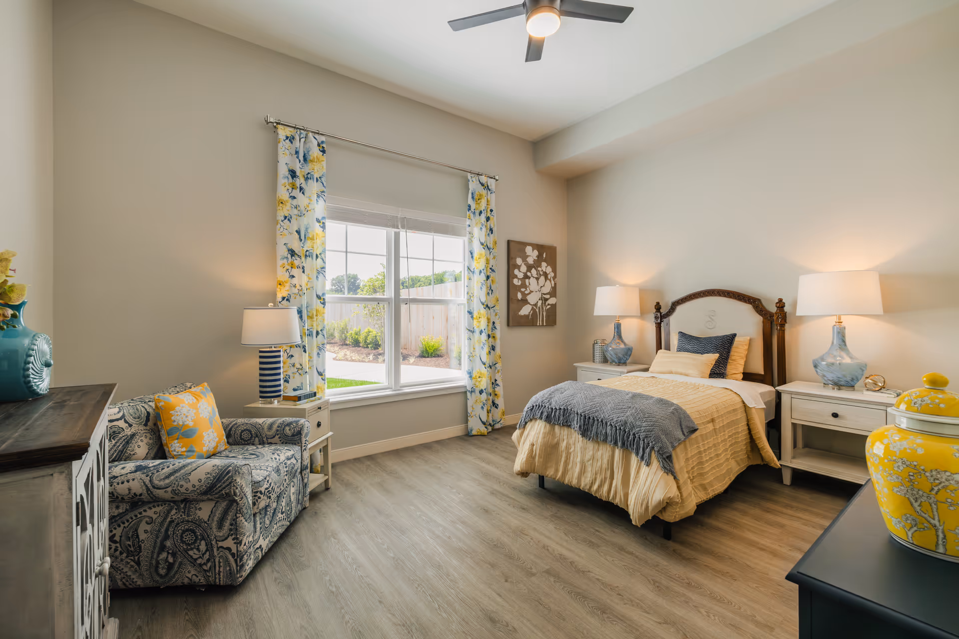 A bright and cozy bedroom with a single bed dressed in yellow and gray bedding, flanked by two white nightstands each with a lamp. A patterned armchair with a yellow floral pillow sits near a window with floral curtains. The room has light-colored walls, wood flooring, and a ceiling fan. Decorative vases and a floral wall art add to the room's decor.