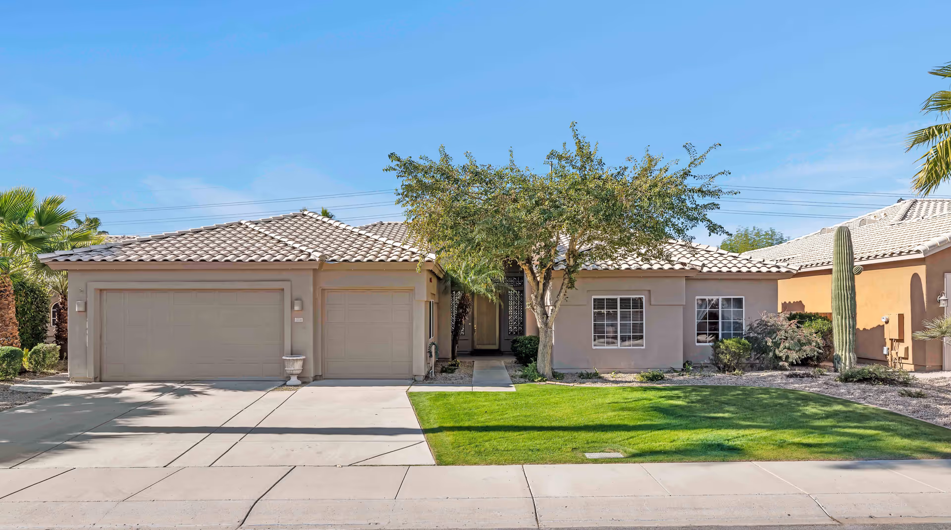 Single-story residential building with a tiled roof, two garage doors, a tree in front, and a well-maintained lawn under a clear blue sky.