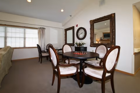 A cozy interior room with a round table surrounded by four upholstered wooden chairs. Behind the table, there is a wall with two large ornate mirrors and a round clock mounted between them. A small cabinet with decorative items is placed beneath the clock. To the left, there is a window with blinds and curtains, and a sofa partially visible. The room has beige carpeting and white walls with recessed lighting in the ceiling.