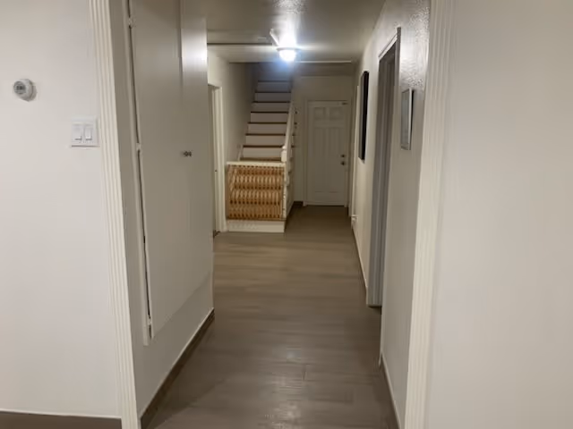 A hallway with light-colored walls and wooden flooring leading to a staircase and a closed door at the end. There are doors and a thermostat on the walls along the hallway.