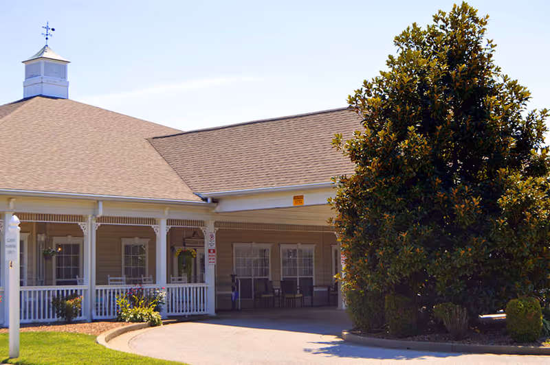 Covered front entrance and porch of a senior living building with a driveway and a large tree in front.