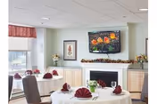 Bright dining room with round tables set with white tablecloths and folded red napkins, chairs, a fireplace and framed artwork on the wall.