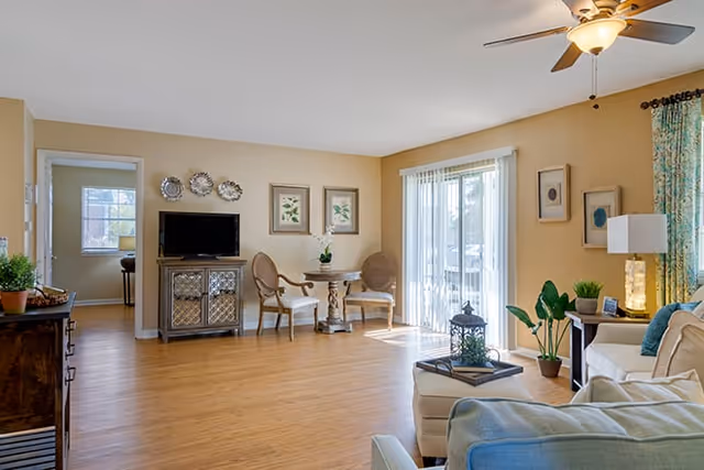 Bright furnished living room with hardwood floors, seating area, TV on a cabinet, small round table with chairs, sliding glass doors and a ceiling fan.