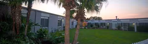 Exterior view of Metairie Health Care Center with palm trees, lawn, and a long low building.
