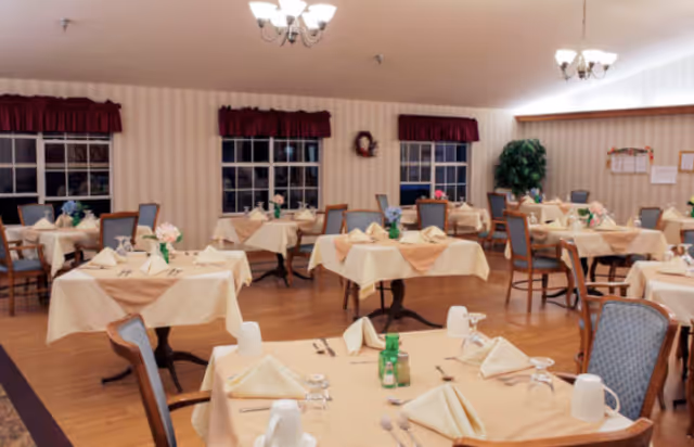 A dining room with multiple tables covered in beige tablecloths and set with napkins, glasses, and silverware. The room has wooden floors, several windows with red valances, and soft lighting from ceiling fixtures. Chairs with blue upholstery surround the tables, and there is a large green plant in the corner.