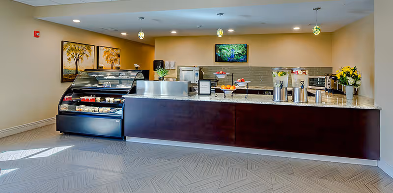 A clean and modern food service counter area with a glass display case containing desserts, a granite countertop with beverage dispensers and fruit bowls, pendant lights hanging from the ceiling, and framed artwork on the wall.