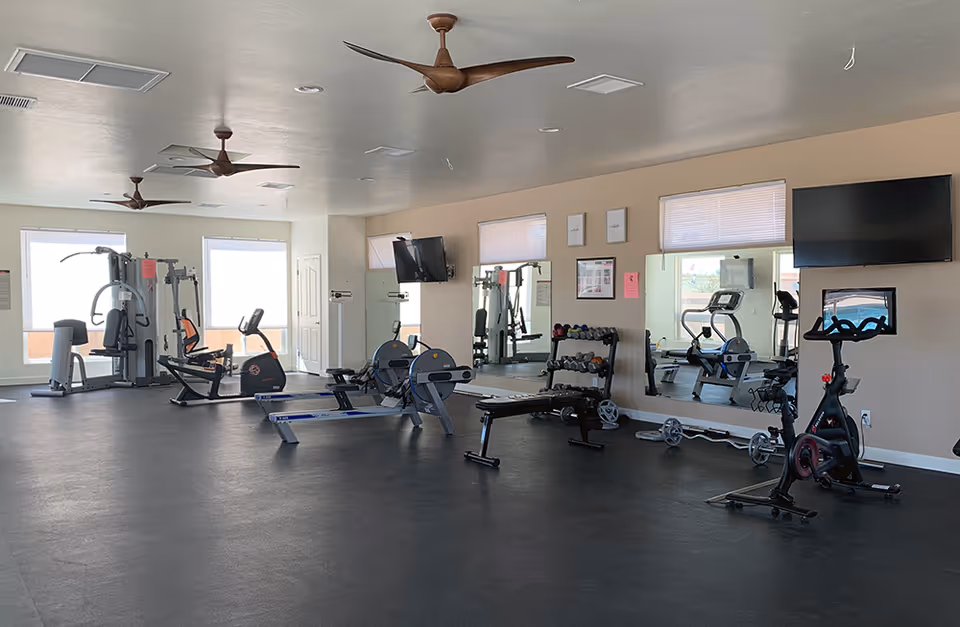 A spacious fitness room with various exercise equipment including stationary bikes, rowing machines, weight bench, dumbbells, and a multi-function gym machine. The room has large windows, ceiling fans, and two wall-mounted televisions above mirrors.