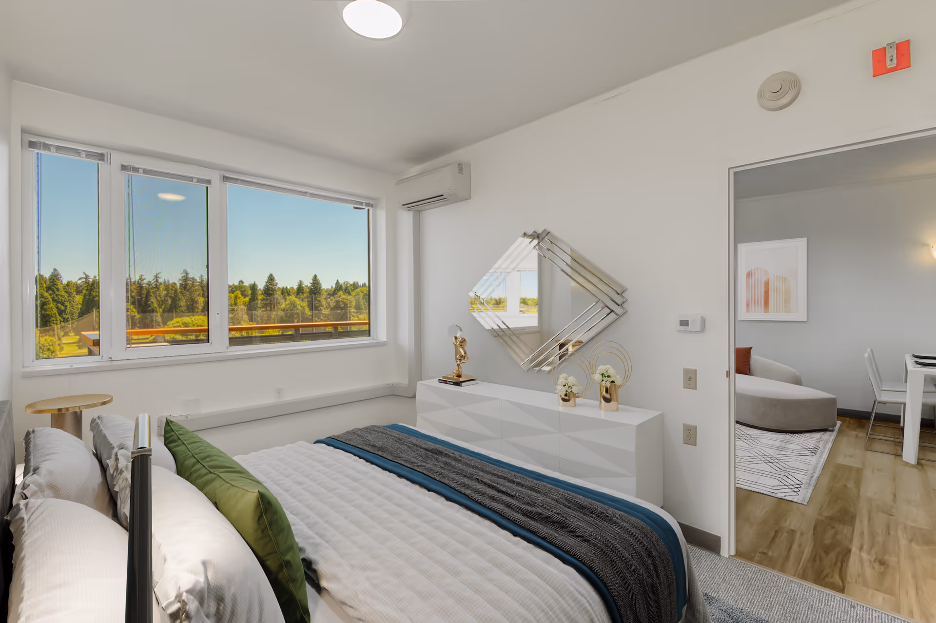 A bright bedroom with a large window showing a view of trees outside. The room features a neatly made bed with white and green pillows, a white dresser with decorative items, and a modern diamond-shaped mirror on the wall. Through an open door, a living area with a sofa and dining table is visible.