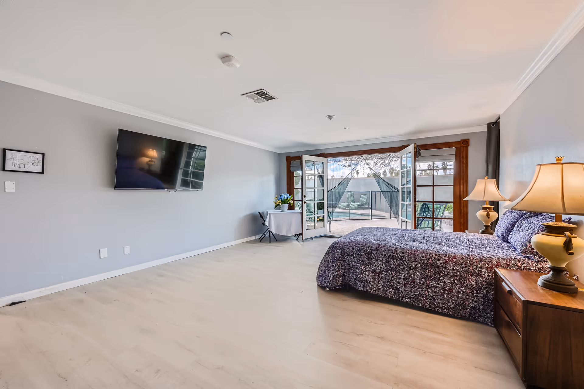 Spacious bedroom with a large bed covered in a purple patterned bedspread, two bedside tables with lamps, a wall-mounted flat screen TV, and French doors opening to an outdoor patio area with seating and a hammock.