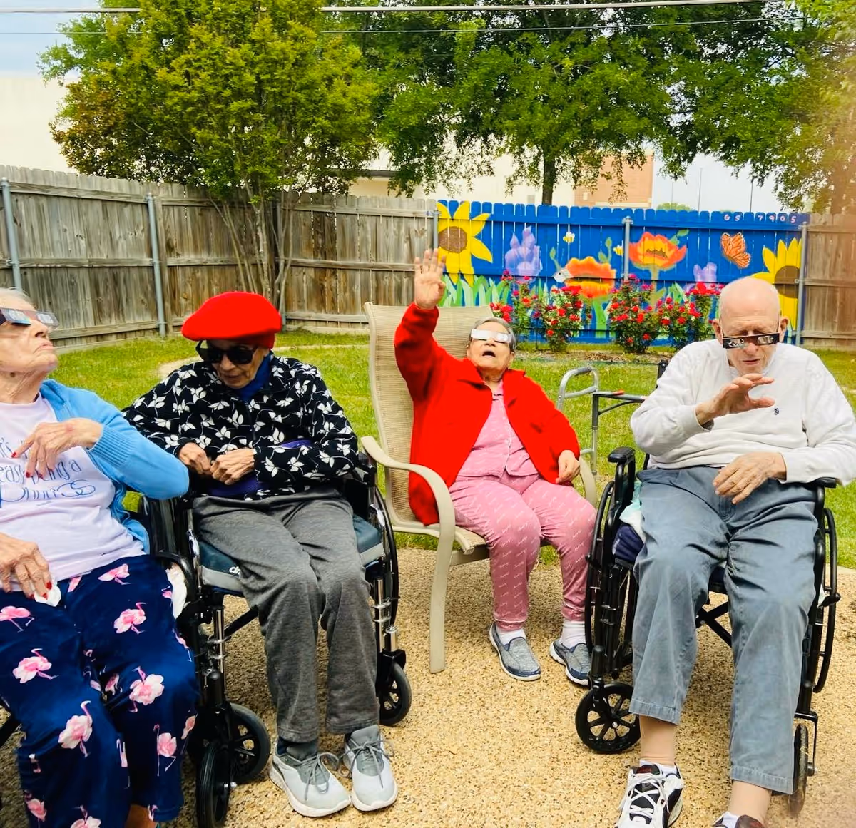 Four elderly individuals sitting outdoors in a garden area with a wooden fence and colorful flower mural in the background. Three of them are in wheelchairs and one is seated in a chair. They are wearing special glasses, possibly for an eclipse or similar event, and appear to be looking up at the sky. The setting is bright and sunny with green grass and trees.